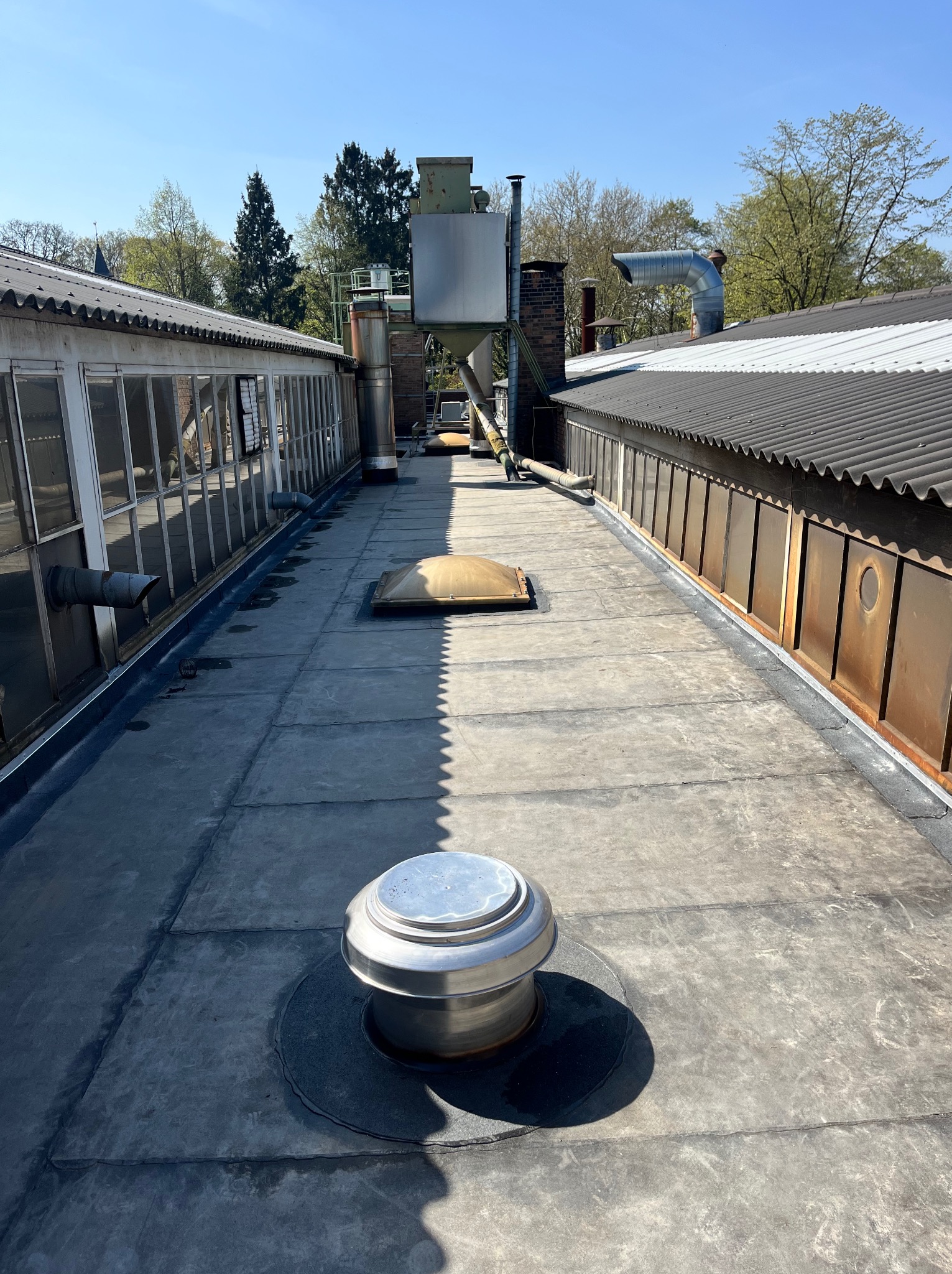 Płaski dach pokryty papą, z metalową wentylacją i świetlikiem. Widoczne kominy, rynny i obróbki blacharskie. W tle drzewa i budynki przemysłowe.
