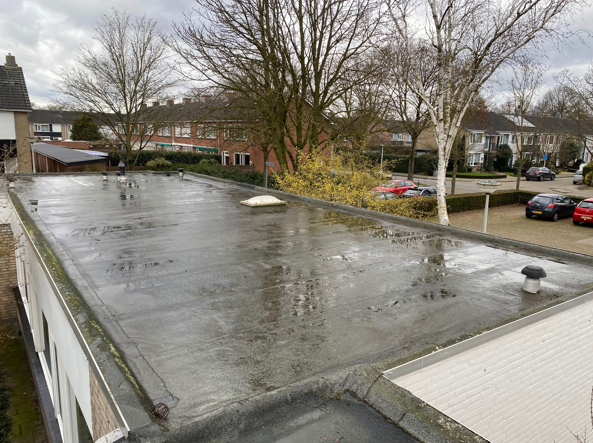 Płaski, mokry dach budynku z widocznymi elementami wentylacyjnymi na tle zabudowy mieszkalnej i drzew. Widok z góry.