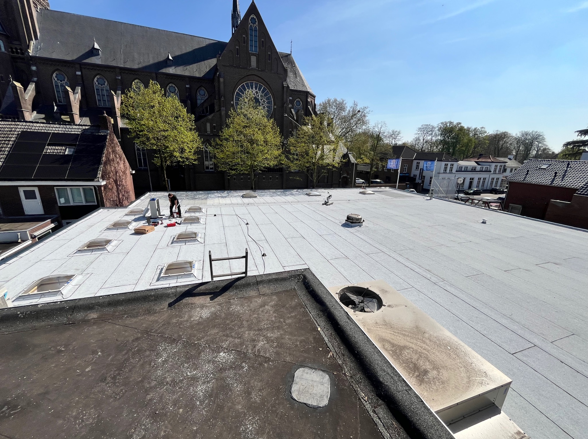 Praca na dachu: dekarz przy świetlikach na jasnoszarym, płaskim dachu z widokiem na kościół w tle. Widoczne narzędzia i materiały dekarskie.