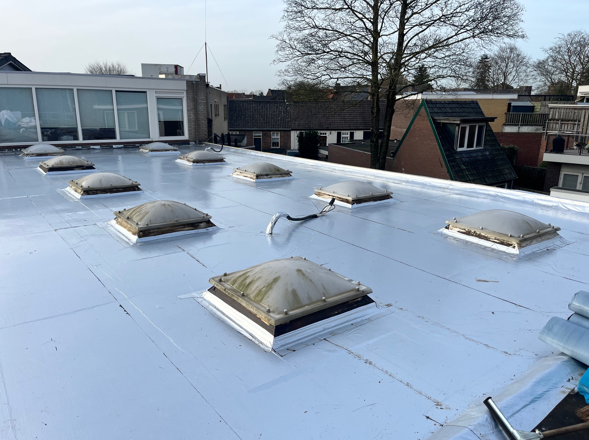 Biały, płaski dach z lukarnami po renowacji, widok z góry. Na dachu widoczne rolki materiału izolacyjnego i narzędzia dekarskie. W tle zabudowa miejska i drzewa.