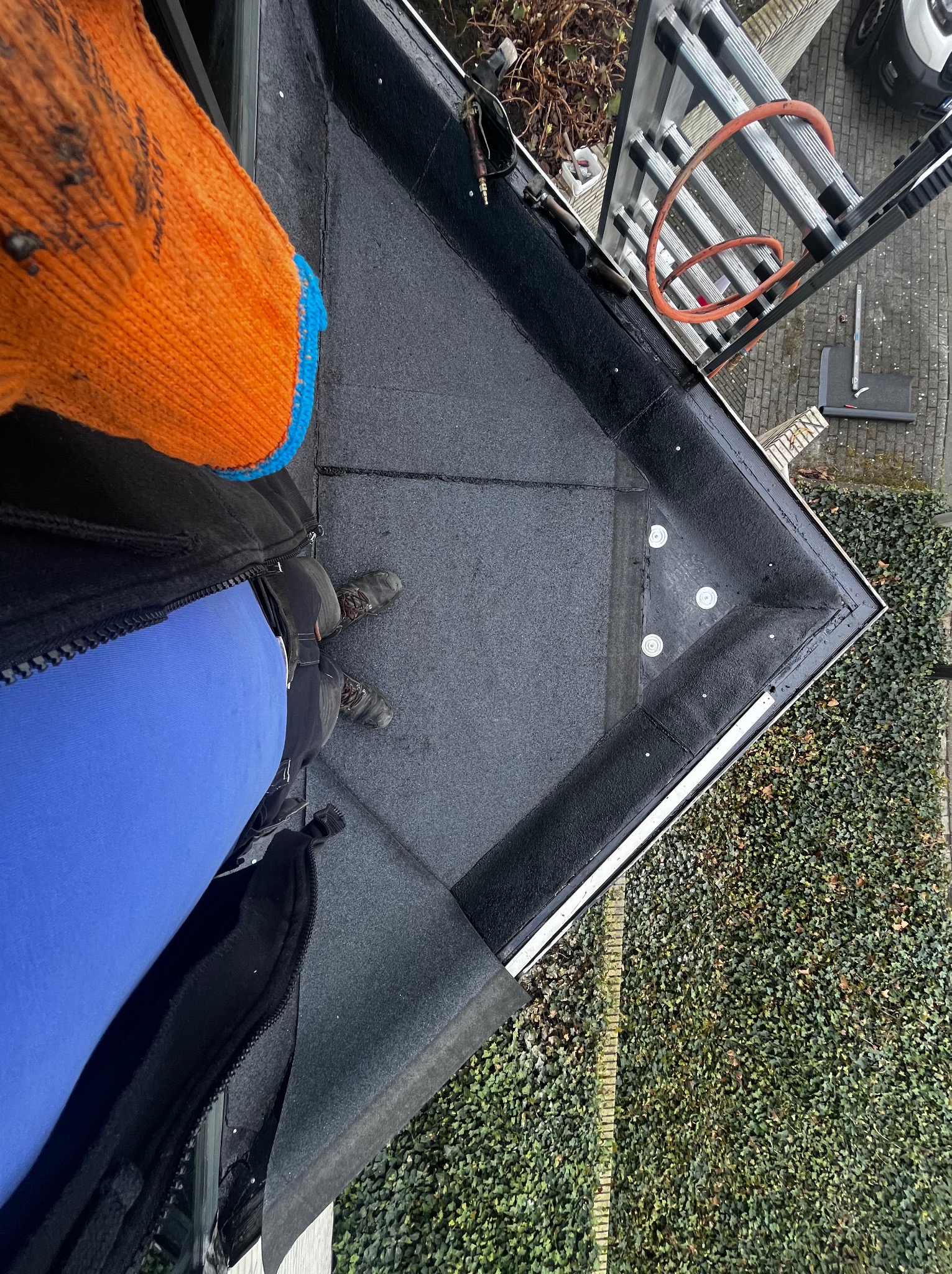 Widok z góry na fragment dachu pokrytego papą, z widoczną drabiną i fragmentem osoby w odzieży roboczej. Dach zabezpieczony, z widocznymi elementami mocującymi.