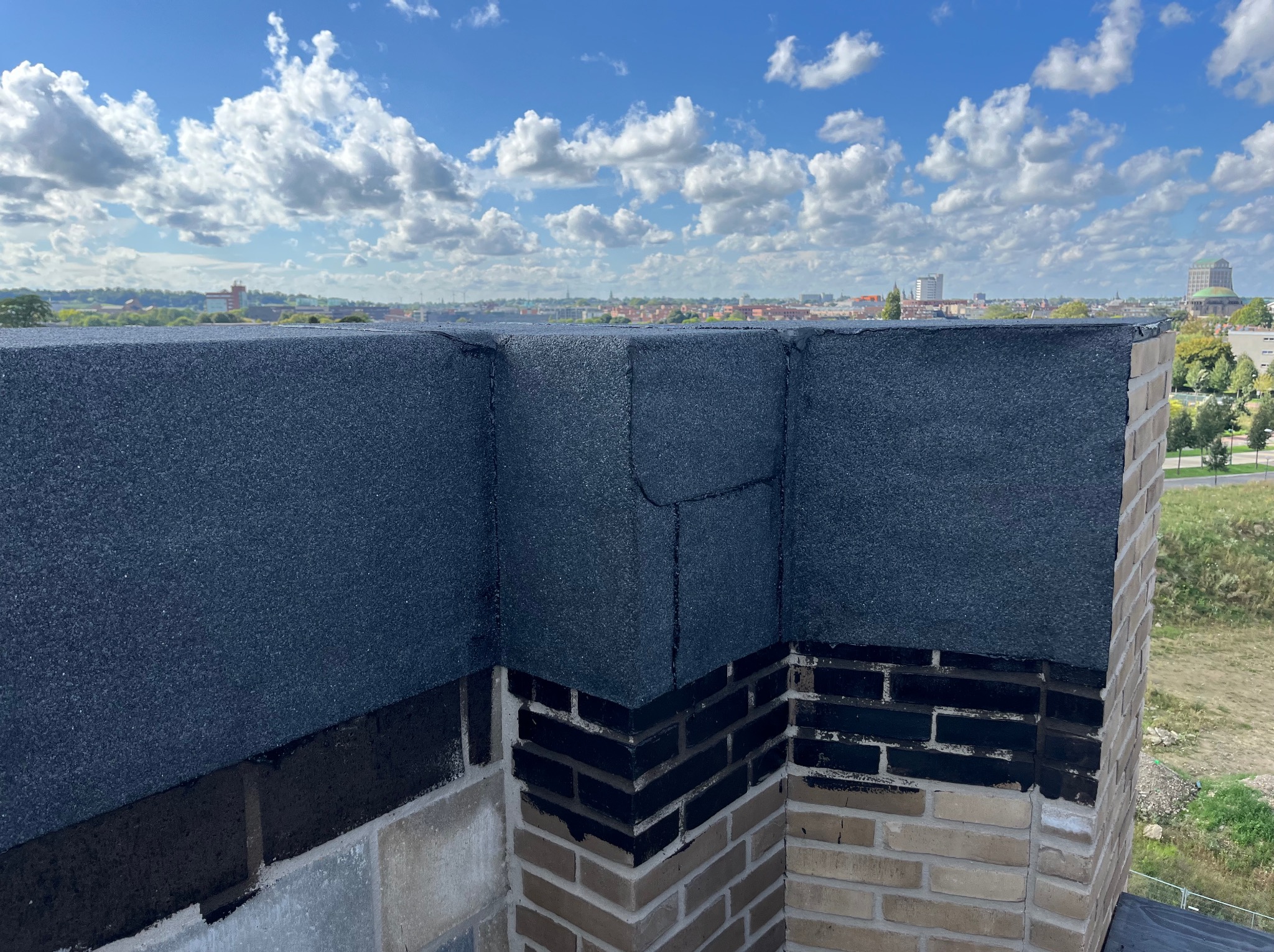 Wykończony fragment dachu płaskiego z papy na murze z cegły, widok na miasto w tle pod błękitnym niebem z chmurami.