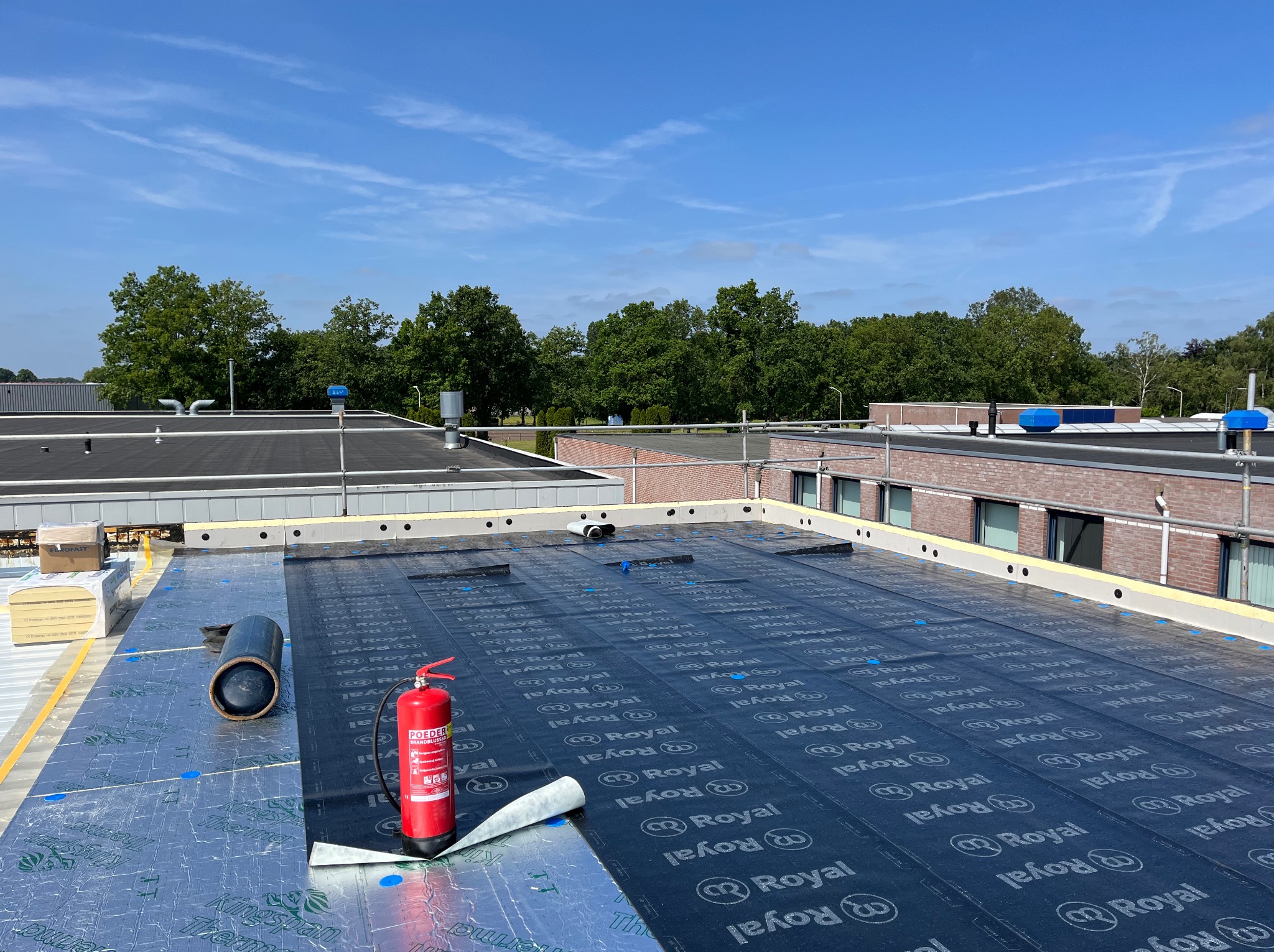 Kładzenie izolacji na dachu płaskim z widocznym sprzętem: butla gazowa i gaśnica. Dach zabezpieczony, niebieskie punkty mocowania. W tle budynki i błękitne niebo.