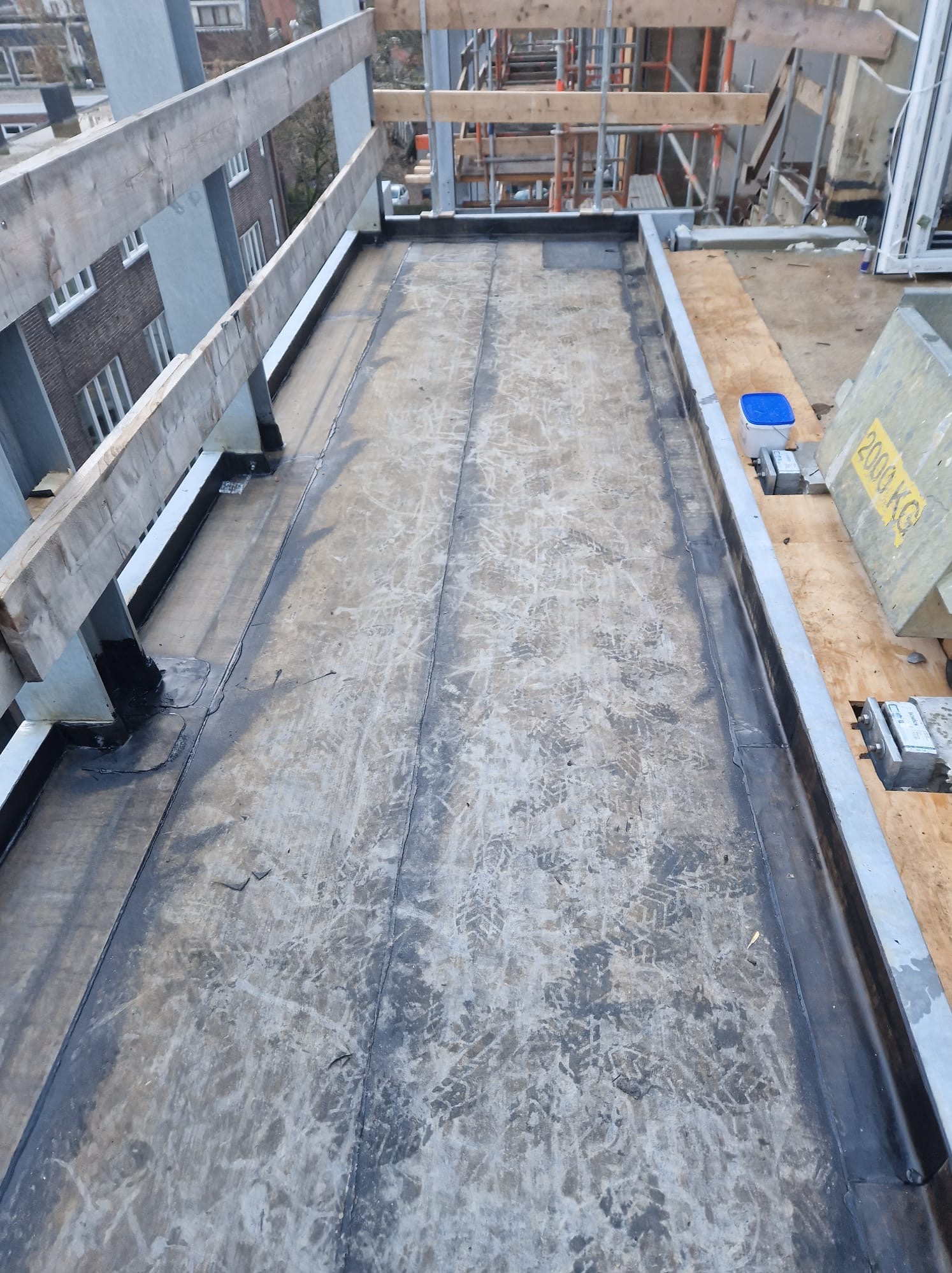 Wykończony płaski dach z papy termozgrzewalnej, zabezpieczony metalową obróbką blacharską i drewnianą barierką. Widoczne ślady robocze i elementy konstrukcyjne.