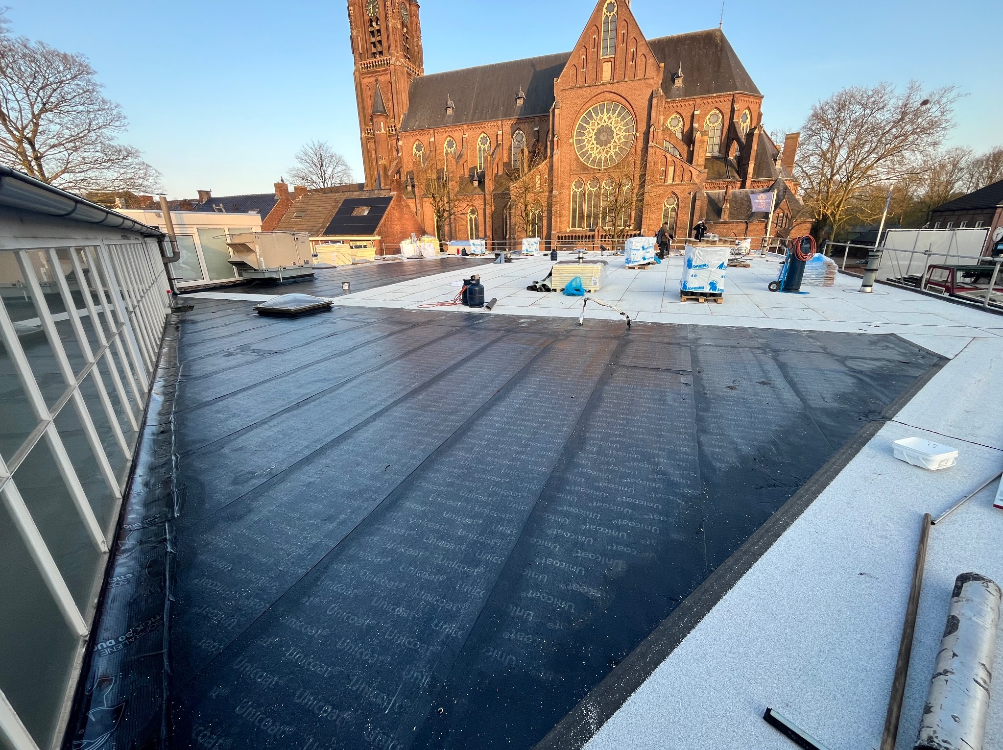 Krycie dachu papą termozgrzewalną Unicoat na tle ceglanej architektury kościoła. Widoczny etap prac z materiałami budowlanymi i narzędziami.