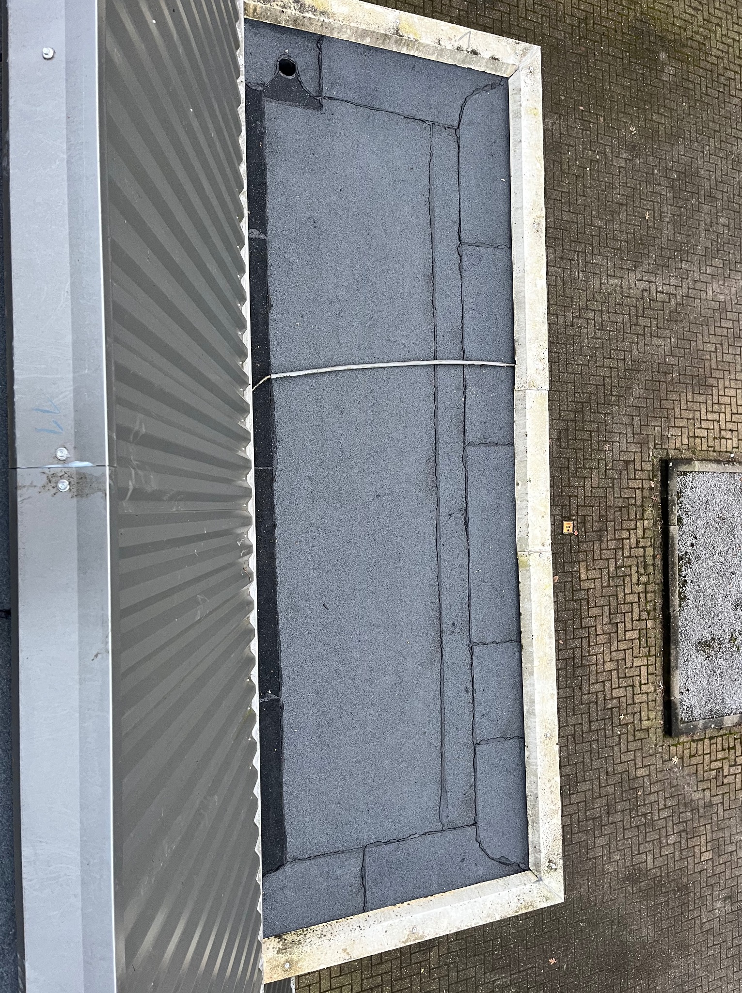 Pionowy widok z góry na fragment dachu pokrytego papą z widocznymi pęknięciami, obok blaszana obróbka i fragment chodnika z kostki. Widoczna instalacja elektryczna.