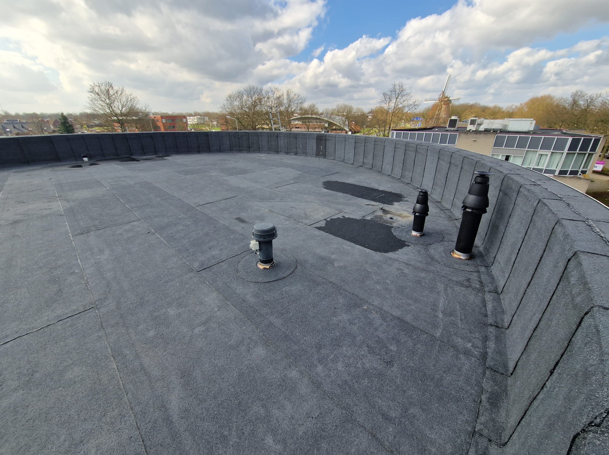 Czarny, płaski dach pokryty papą z widocznymi kominkami wentylacyjnymi i obróbkami blacharskimi. W tle budynki i wiatrak.