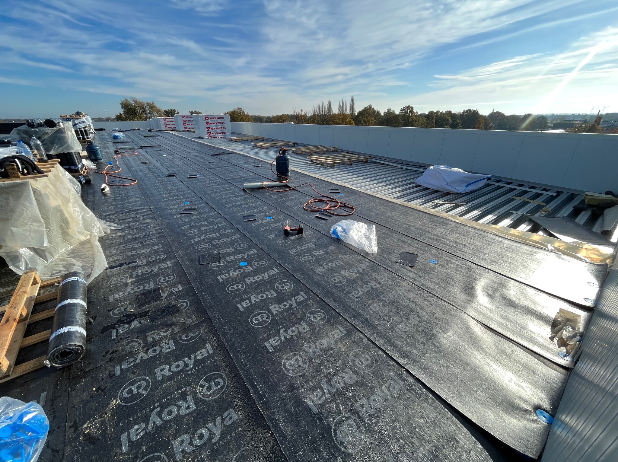 Dach pokryty membraną bitumiczną marki Royal. Widoczne rolki membrany, palety z materiałami izolacyjnymi Rockwool i narzędzia dekarskie. W tle błękitne niebo.