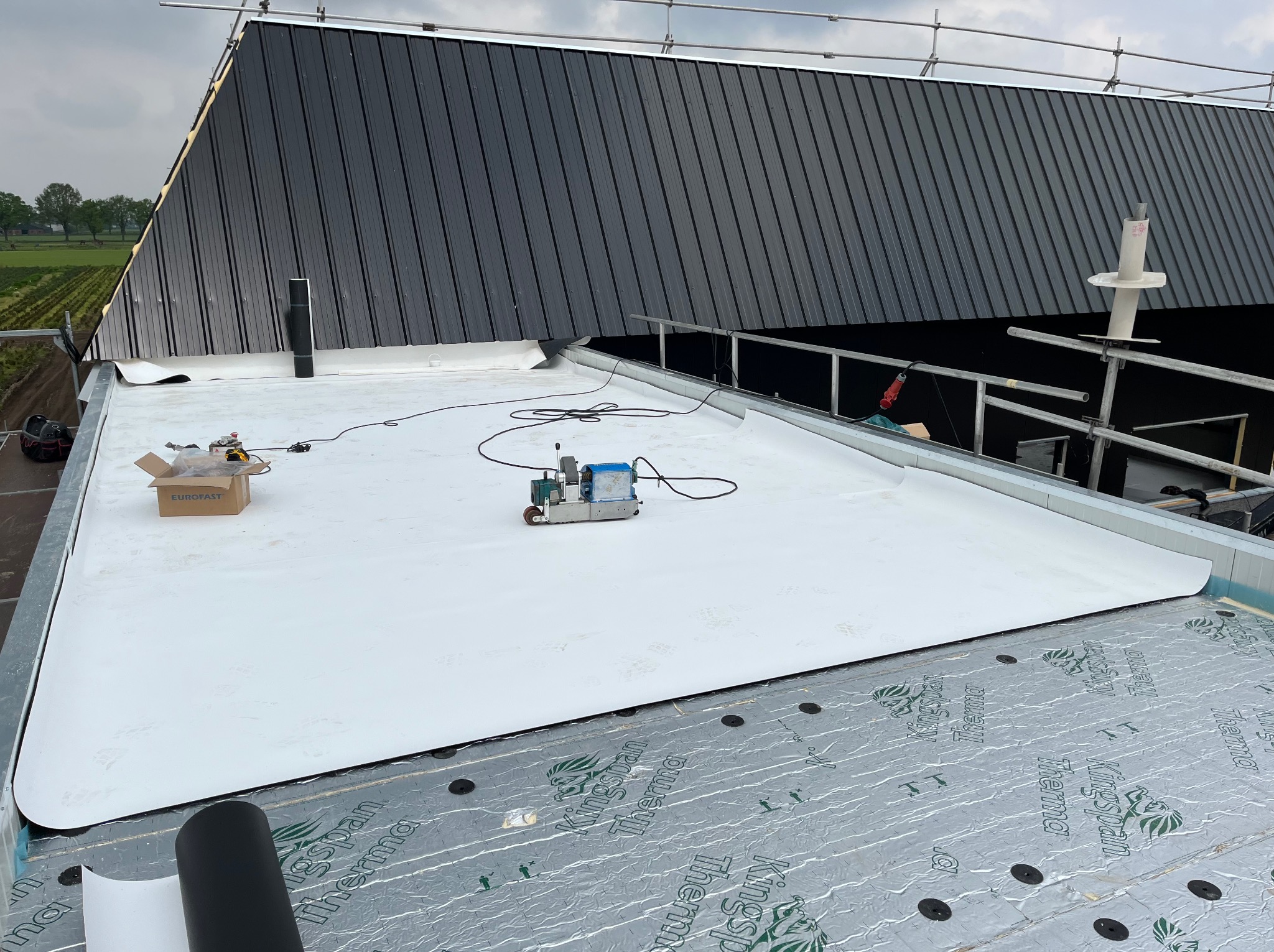 Biała membrana dachowa na izolacji Kingspan Therma, zgrzewarka w użyciu, widoczny fragment czarnego dachu z blachy trapezowej. Prace dekarskie w toku.