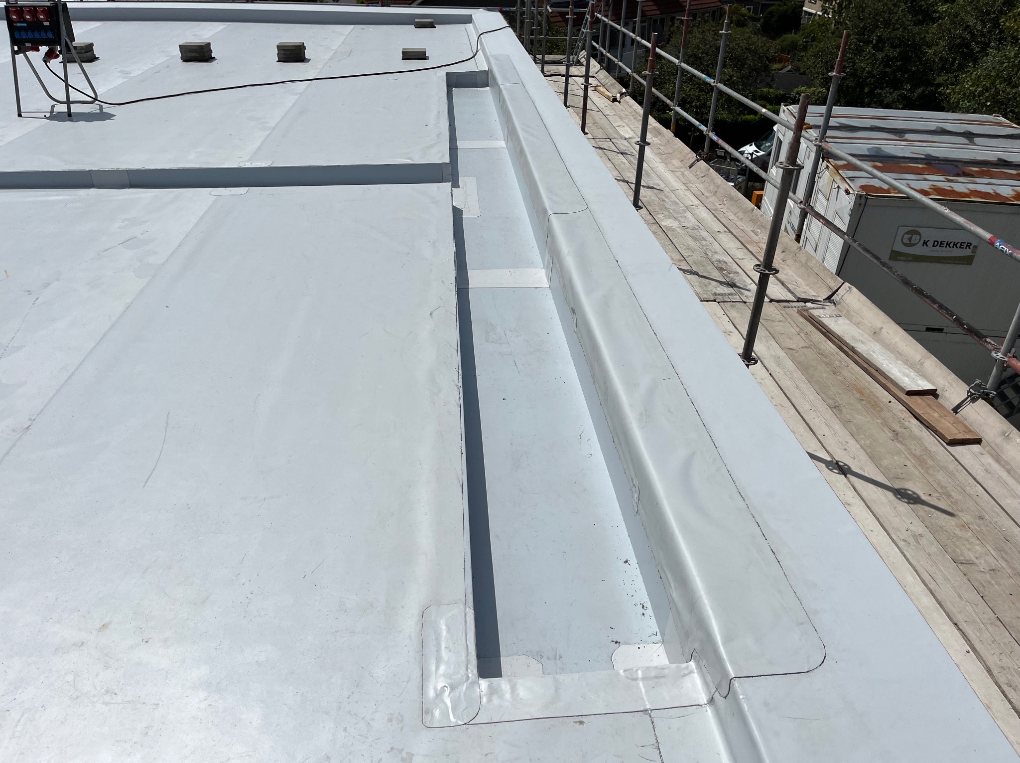 Szary, płaski dach pokryty membraną, z widocznymi obróbkami blacharskimi i elementami instalacji. Ujęcie z rusztowania, z kontenerem w tle.