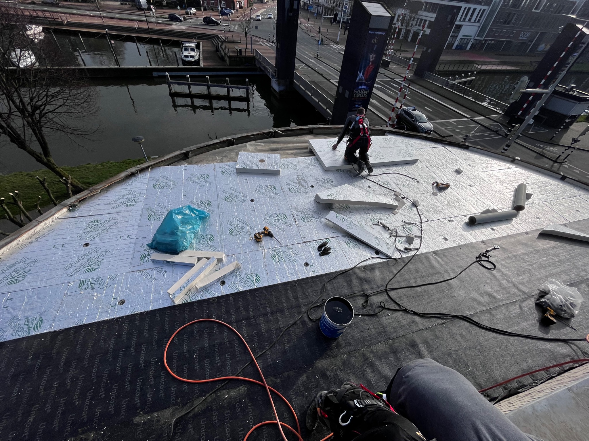 Prace dekarskie na dachu budynku z widokiem na kanał i most zwodzony. Izolacja termiczna Kingspan Thermaroof, narzędzia pracy. Dekarz w uprzęży zabezpieczającej.