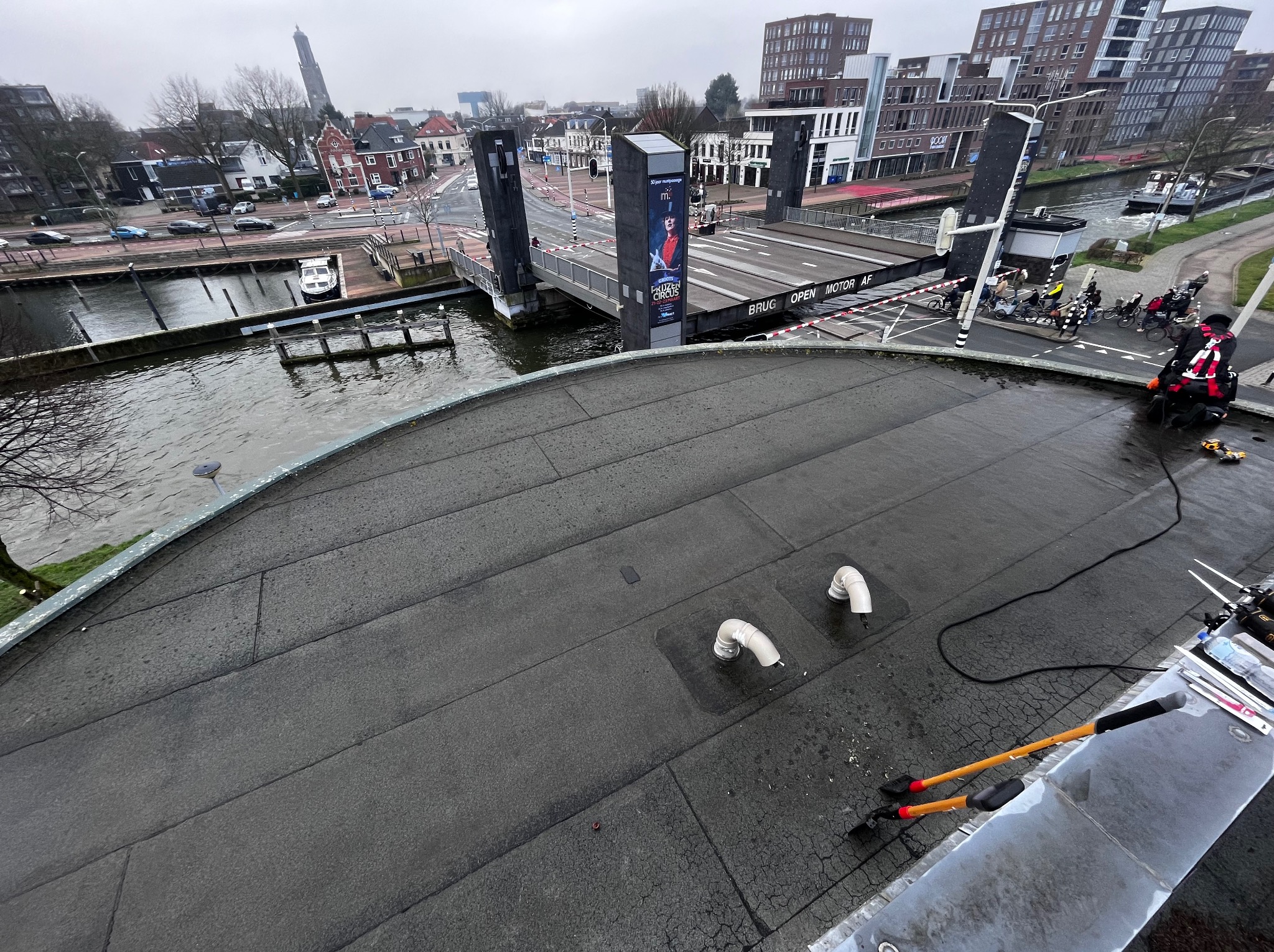 Widok z góry na płaski dach z papy na tle miasta z kanałem i mostem zwodzonym. Widoczne narzędzia dekarskie i elementy wentylacji. Pochmurny dzień.