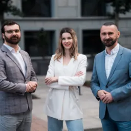 Zespół agencji nieruchomości: kobieta w bieli i dwaj mężczyźni w marynarkach prezentują się na tle nowoczesnego budynku.