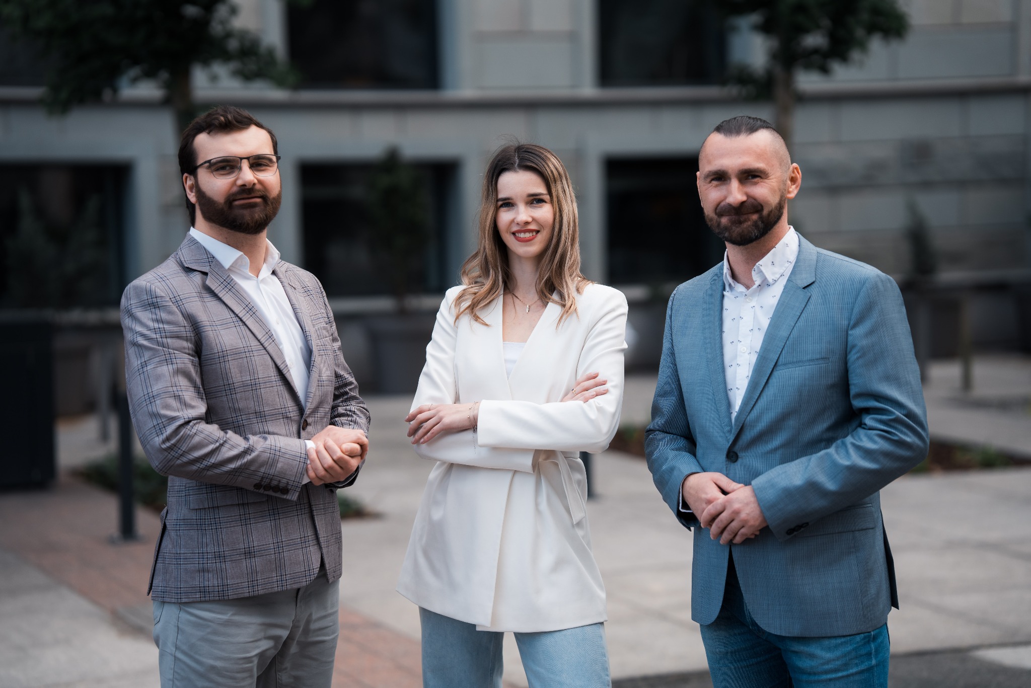 Zespół agencji nieruchomości: kobieta w bieli i dwaj mężczyźni w marynarkach prezentują się na tle nowoczesnego budynku.