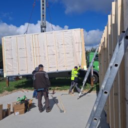 TWÓJ DOM EXPERT SPÓŁKA Z OGRANICZONĄ ODPOWIEDZIALNOŚCIĄ - Montaż ściany szkieletowej domu z użyciem dźwigu. Widoczni pracownicy w kamizelkach, drabina oparta o ścianę i przygotowane elementy konstrukcyjne.