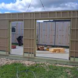 TWÓJ DOM EXPERT SPÓŁKA Z OGRANICZONĄ ODPOWIEDZIALNOŚCIĄ - Montaż ściany domu szkieletowego z użyciem dźwigu, widoczna drabina oparta o ścianę, w tle las i pracownik z narzędziem Hilti.