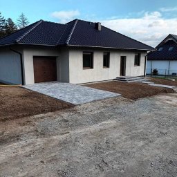 TWÓJ DOM EXPERT SPÓŁKA Z OGRANICZONĄ ODPOWIEDZIALNOŚCIĄ - Parterowy dom z garażem i brukowanym podjazdem. Nowoczesna architektura, szara elewacja, ciemna dachówka. Uporządkowany teren wokół domu.