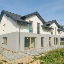 TWÓJ DOM EXPERT SPÓŁKA Z OGRANICZONĄ ODPOWIEDZIALNOŚCIĄ - Nowoczesne domy w zabudowie szeregowej z dwukolorową elewacją (szary dół, biała góra) i ciemną dachówką, widok z zewnątrz, kostka brukowa na podejściu, trawa.