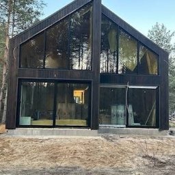 TWÓJ DOM EXPERT SPÓŁKA Z OGRANICZONĄ ODPOWIEDZIALNOŚCIĄ - Nowoczesny dom z ciemnego drewna z dużymi przeszkleniami, widoczne drzewa w tle. Surowy teren przed budynkiem, świeżo wylany fundament.