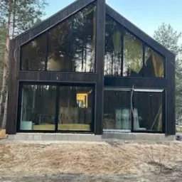 Nowoczesny dom z ciemnego drewna z dużymi przeszkleniami, widoczne drzewa w tle. Surowy teren przed budynkiem, świeżo wylany fundament.