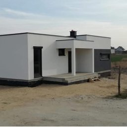 TWÓJ DOM EXPERT SPÓŁKA Z OGRANICZONĄ ODPOWIEDZIALNOŚCIĄ - Nowoczesny, parterowy dom z płaskim dachem, wykończony w bieli i szarości, z widocznym tarasem wejściowym i kominem. W tle zabudowa jednorodzinna i pole.