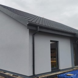 TWÓJ DOM EXPERT SPÓŁKA Z OGRANICZONĄ ODPOWIEDZIALNOŚCIĄ - Nowoczesny dom z szarą elewacją i ciemną dachówką, widok z boku. Uwagę zwracają czarne rynny i okna. Dom w trakcie wykończenia, widoczne elementy fundamentu.