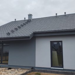 TWÓJ DOM EXPERT SPÓŁKA Z OGRANICZONĄ ODPOWIEDZIALNOŚCIĄ - Wykończony dom z szarą dachówką i elewacją w dwóch odcieniach szarości, z widocznymi oknami i fragmentem ogrodu w pochmurny dzień. Detal dachu z kominkiem.