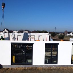 TWÓJ DOM EXPERT SPÓŁKA Z OGRANICZONĄ ODPOWIEDZIALNOŚCIĄ - Montaż modułu domu z oknami przez dźwig na tle zabudowy jednorodzinnej i błękitnego nieba. Widoczni pracownicy w odblaskowych kamizelkach.