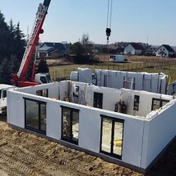 TWÓJ DOM EXPERT SPÓŁKA Z OGRANICZONĄ ODPOWIEDZIALNOŚCIĄ - Montaż domu modułowego z użyciem dźwigu. Widoczne elementy konstrukcyjne, okna i pracownik wewnątrz. W tle zabudowa jednorodzinna i drzewa.