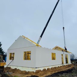 TWÓJ DOM EXPERT SPÓŁKA Z OGRANICZONĄ ODPOWIEDZIALNOŚCIĄ - Montaż domu modułowego z użyciem dźwigu. Białe ściany z otworami okiennymi, drewniana konstrukcja dachu w trakcie budowy. Na zewnątrz widoczny krajobraz.