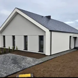 Nowoczesny dom z czarną dachówką i minimalistyczną bryłą w Poznaniu. Widoczne detale wykończenia elewacji i otoczenia budynku. Charakterystyczne, wąskie okna.