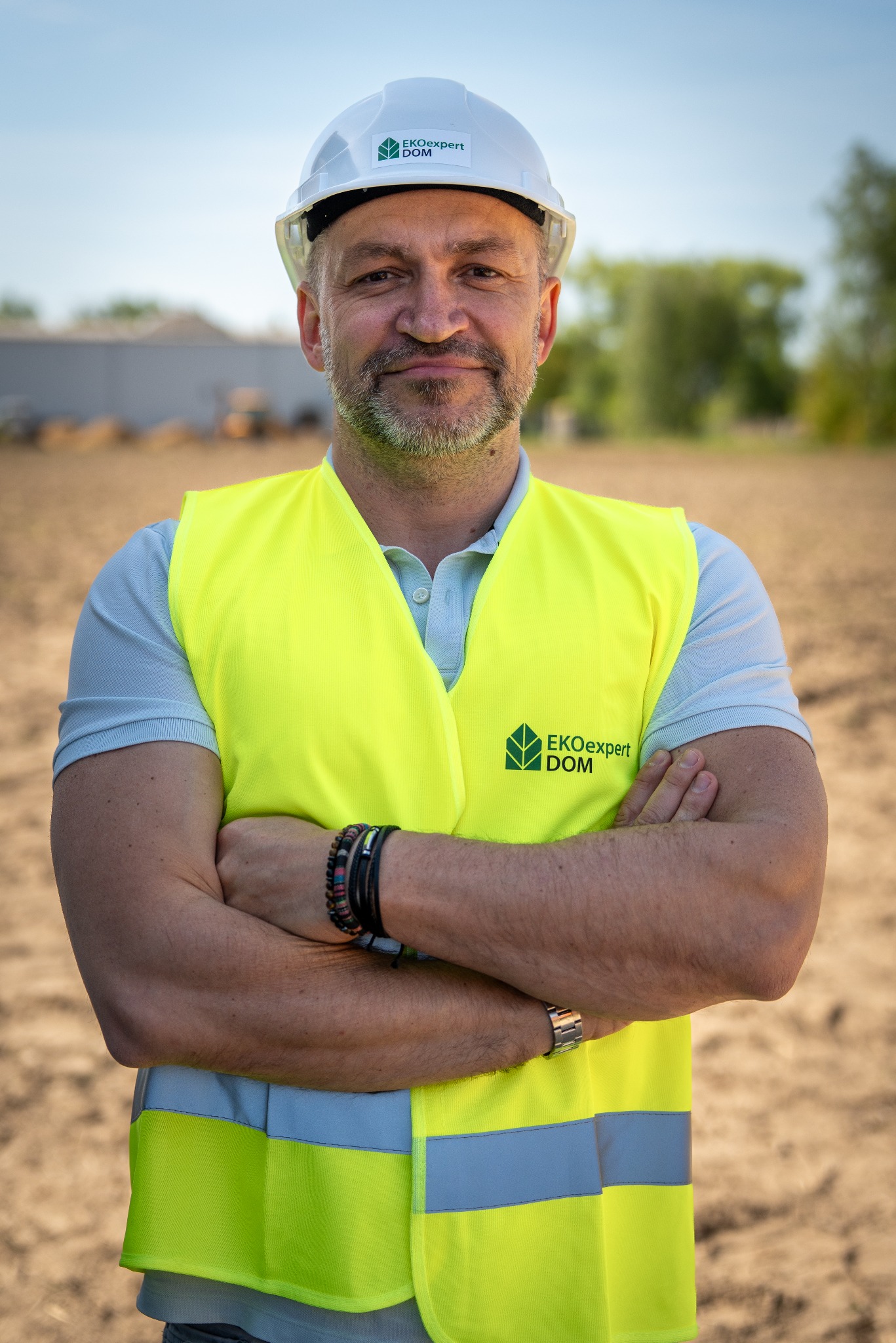 Portret inżyniera w kasku i kamizelce EKOexpert DOM na tle placu budowy. Uśmiechnięty mężczyzna z brodą, skrzyżowane ręce, naturalne światło.