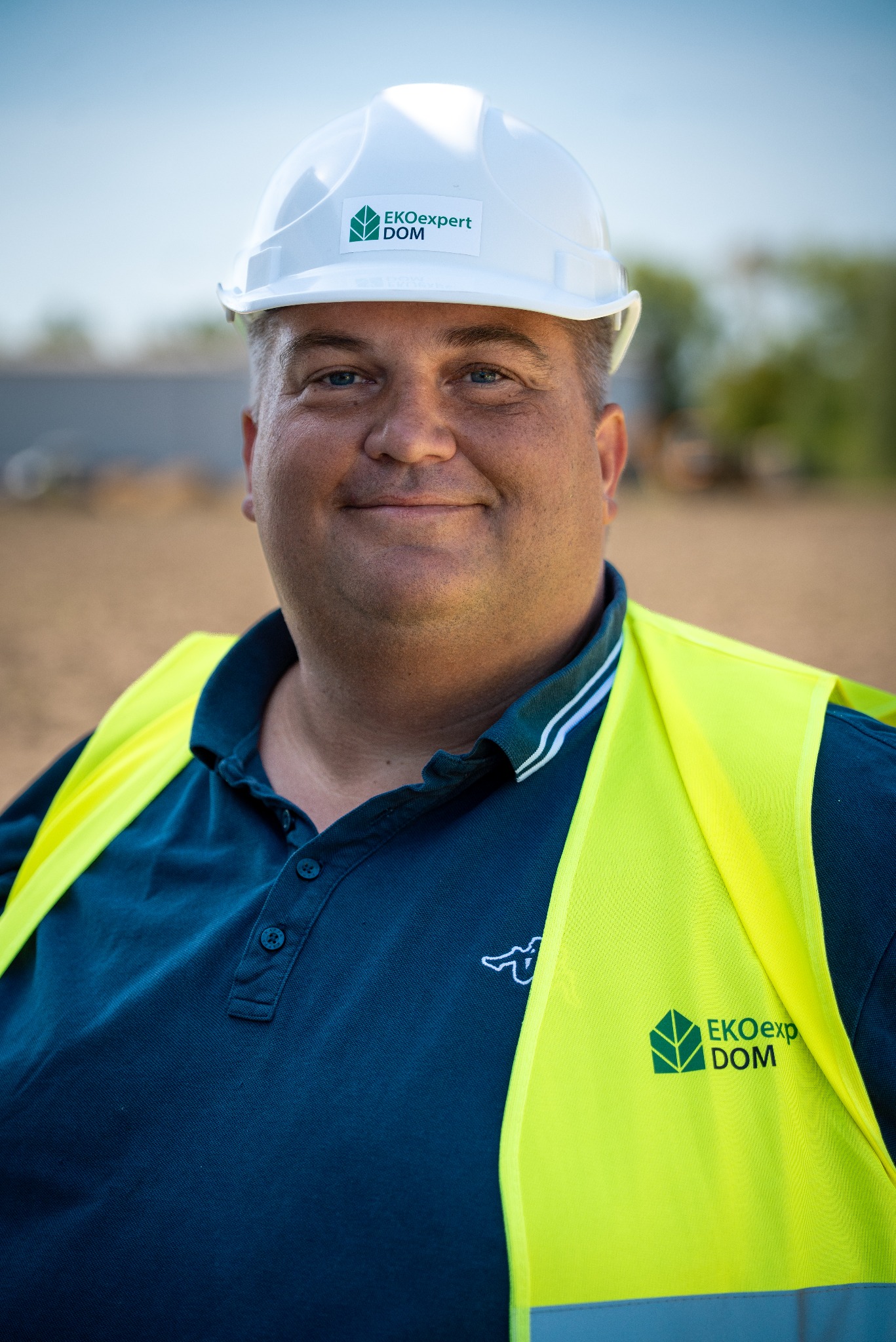 Portret uśmiechniętego inżyniera w kasku z logo EkoExpert Dom i żółtej kamizelce odblaskowej na tle placu budowy.