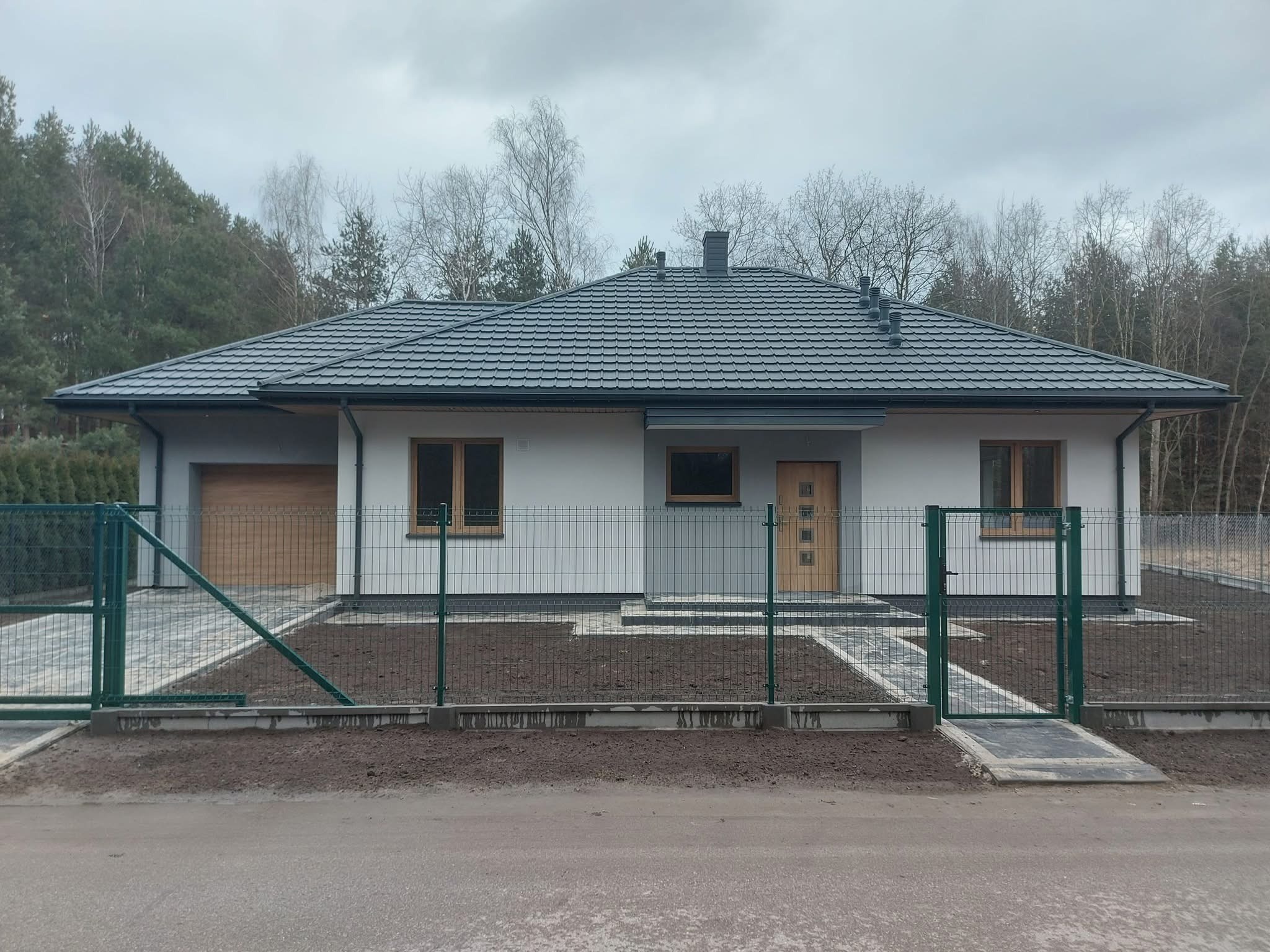 Parterowy dom z garażem w Poznaniu, wykończony w jasnych barwach z szarą dachówką. Widoczny fragment ogrodzenia z siatki i utwardzony podjazd.