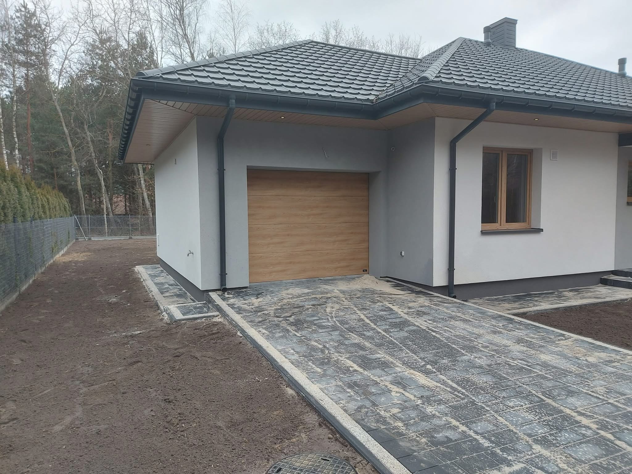 Nowoczesny dom z garażem i brukowanym podjazdem w trakcie wykańczania. Widoczne szare ściany, drewniana brama garażowa i ciemna dachówka. Tło stanowią drzewa.
