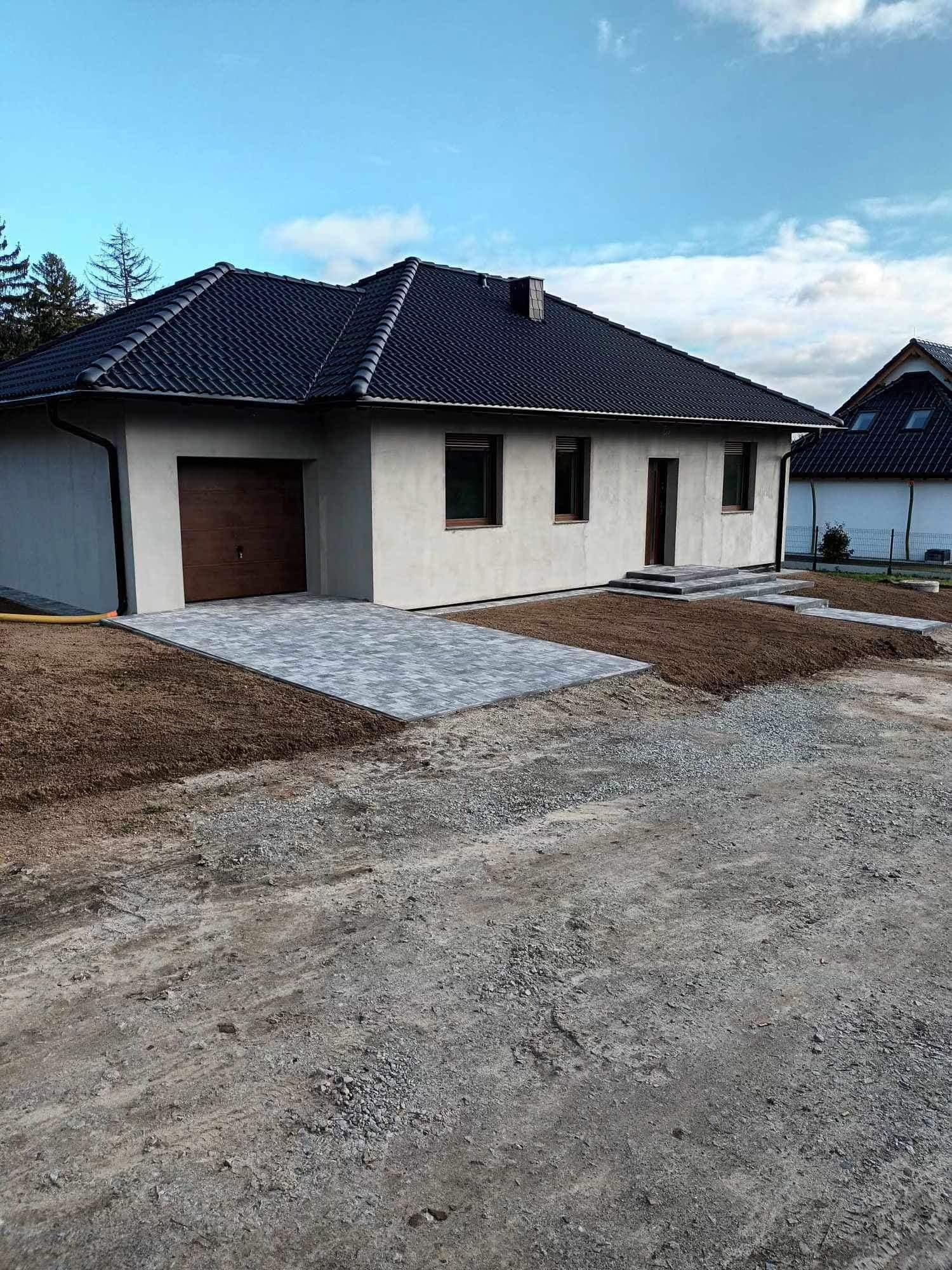 Parterowy dom z garażem i brukowanym podjazdem. Nowoczesna architektura, szara elewacja, ciemna dachówka. Uporządkowany teren wokół domu.