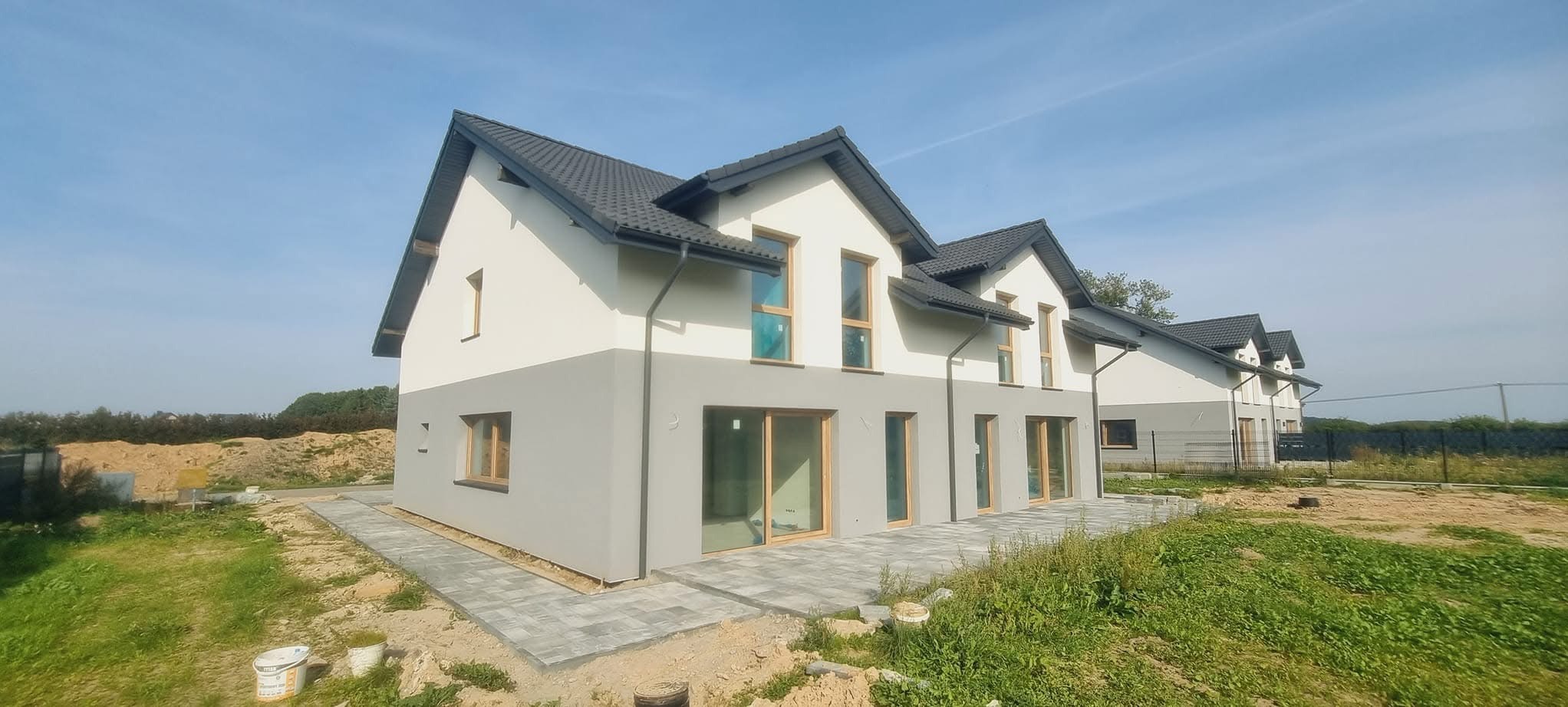 Nowoczesne domy w zabudowie szeregowej z dwukolorową elewacją (szary dół, biała góra) i ciemną dachówką, widok z zewnątrz, kostka brukowa na podejściu, trawa.