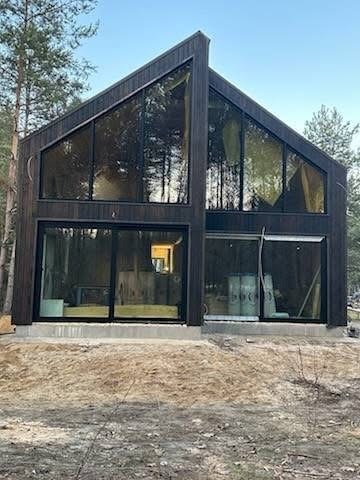 Nowoczesny dom z ciemnego drewna z dużymi przeszkleniami, widoczne drzewa w tle. Surowy teren przed budynkiem, świeżo wylany fundament.