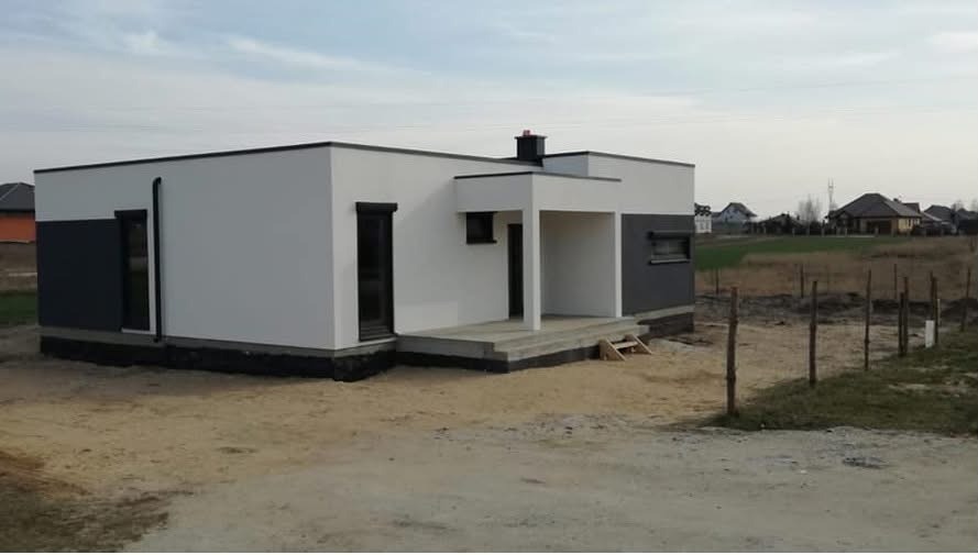 Nowoczesny, parterowy dom z płaskim dachem, wykończony w bieli i szarości, z widocznym tarasem wejściowym i kominem. W tle zabudowa jednorodzinna i pole.