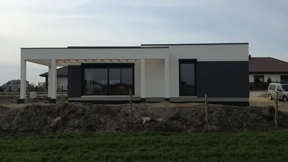 Nowoczesny, parterowy dom z płaskim dachem w biało-szarej kolorystyce. Widoczny taras zadaszony pergolą oraz duże okna. Dom w trakcie wykończenia, otoczony ziemią.