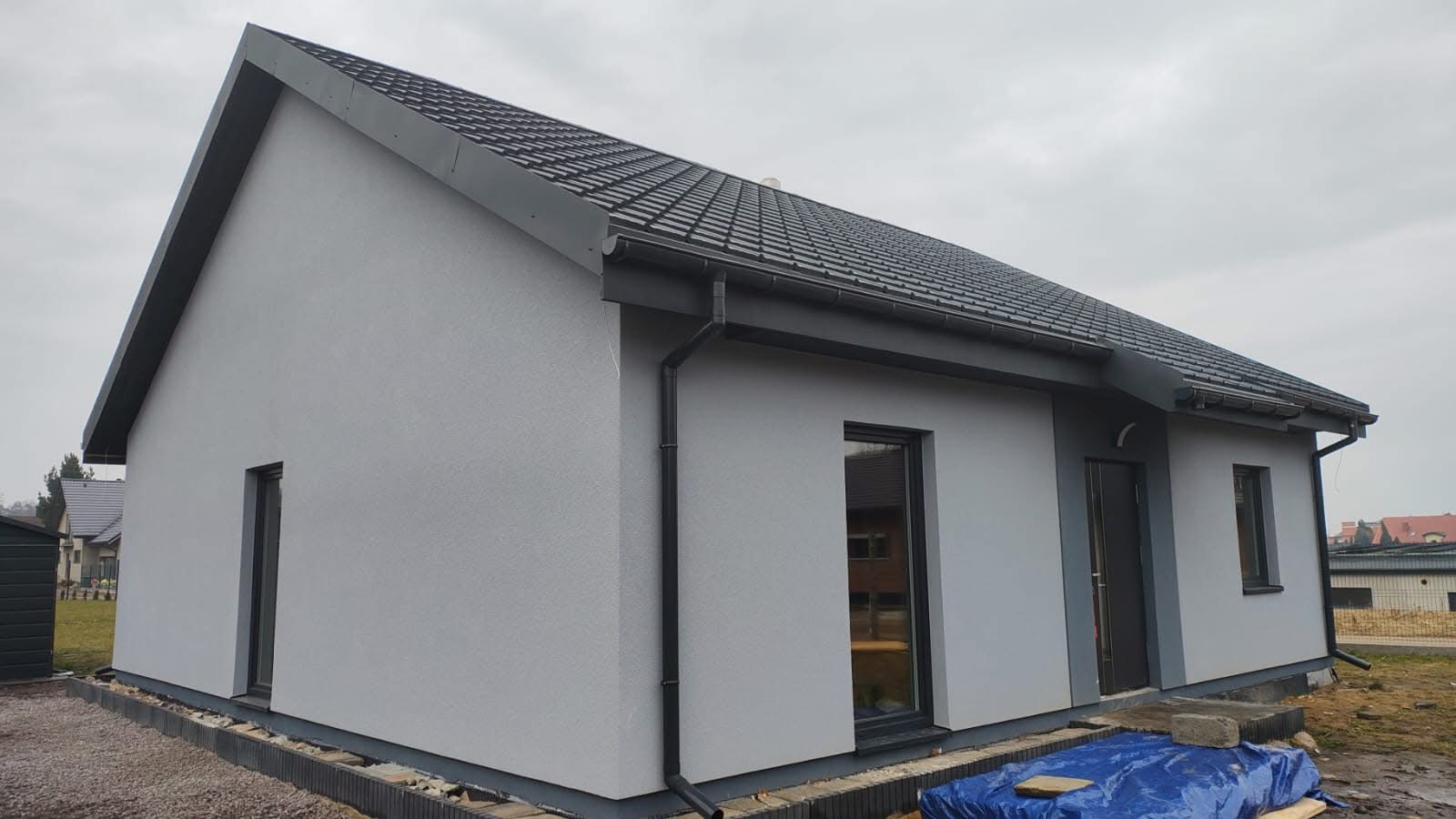 Nowoczesny dom z szarą elewacją i ciemną dachówką, widok z boku. Uwagę zwracają czarne rynny i okna. Dom w trakcie wykończenia, widoczne elementy fundamentu.