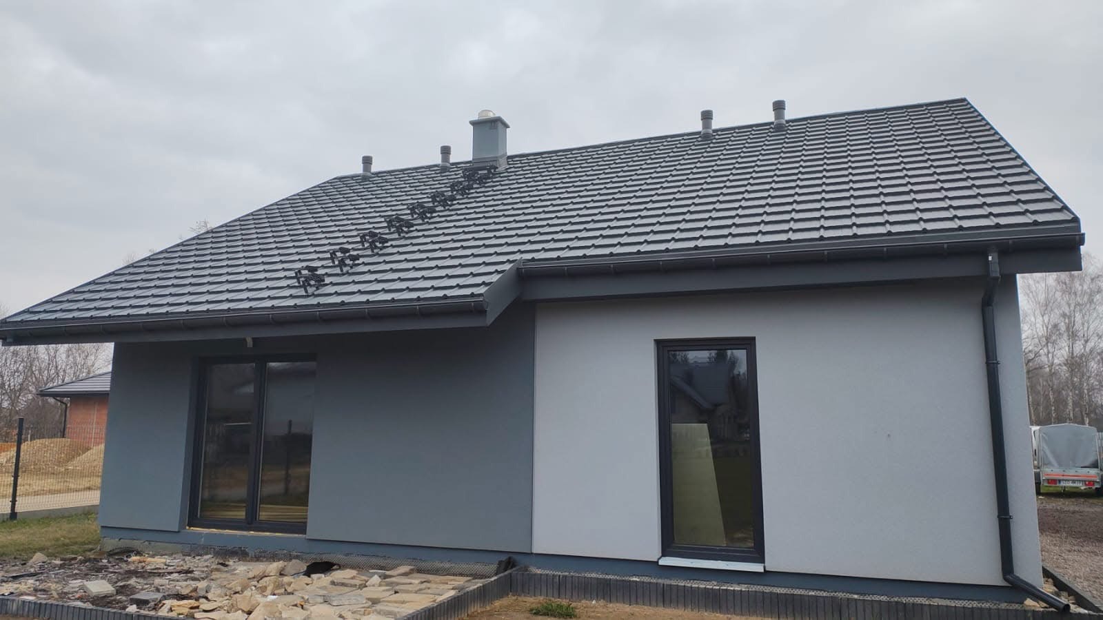 Wykończony dom z szarą dachówką i elewacją w dwóch odcieniach szarości, z widocznymi oknami i fragmentem ogrodu w pochmurny dzień. Detal dachu z kominkiem.