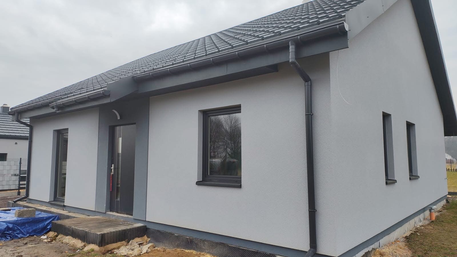 Parterowy dom z szarą elewacją, ciemną dachówką i czarnymi rynnami. Widoczne okna i drzwi wejściowe. Częściowo zagospodarowany teren wokół budynku.