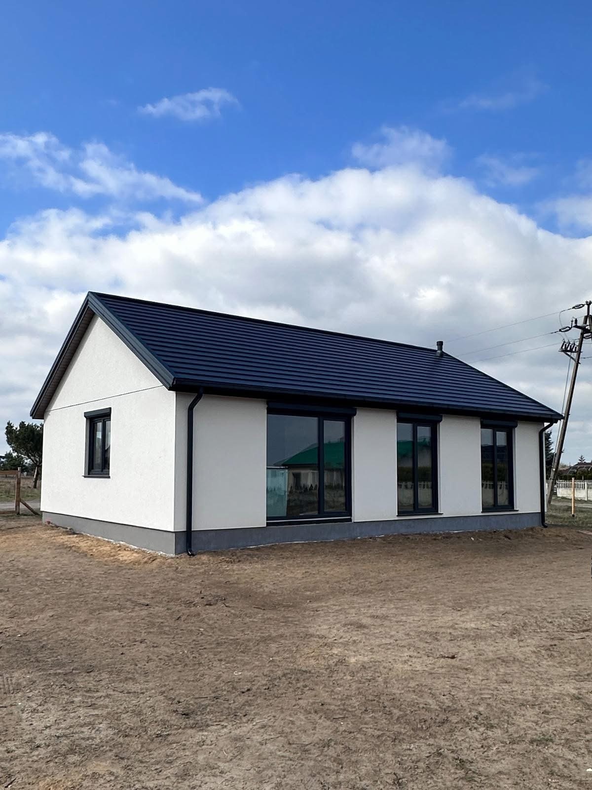 Parterowy dom z ciemną dachówką i dużymi oknami, w trakcie wykończenia terenu, na tle błękitnego nieba z chmurami. Widok z boku, z zewnątrz.