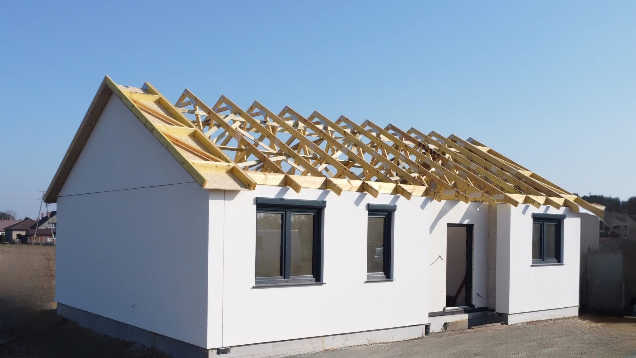 Surowy stan domu z białymi ścianami i drewnianą konstrukcją dachu w trakcie budowy, okna w kolorze antracytowym, błękitne niebo w tle.