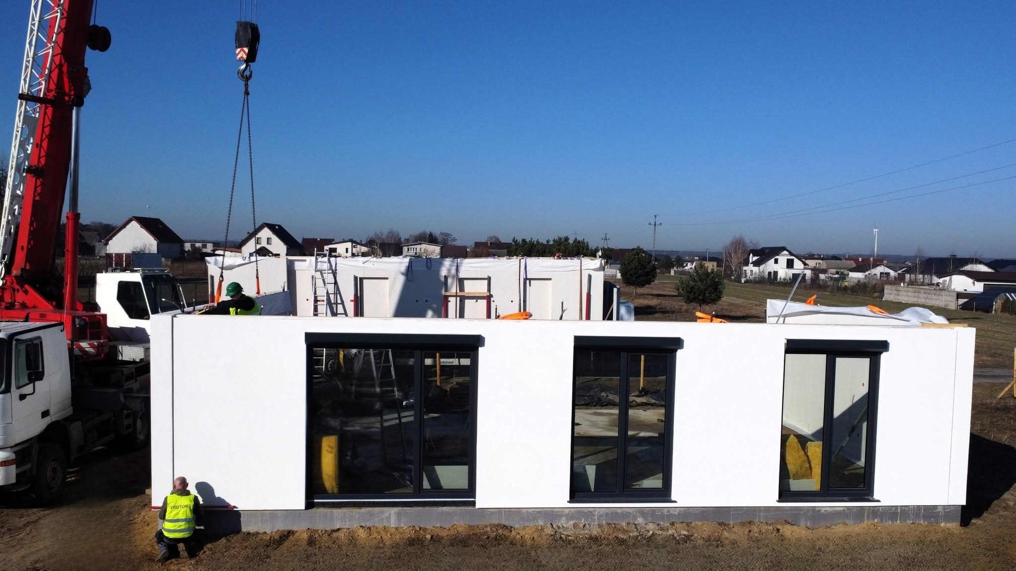 Montaż modułu domu z oknami przez dźwig na tle zabudowy jednorodzinnej i błękitnego nieba. Widoczni pracownicy w odblaskowych kamizelkach.