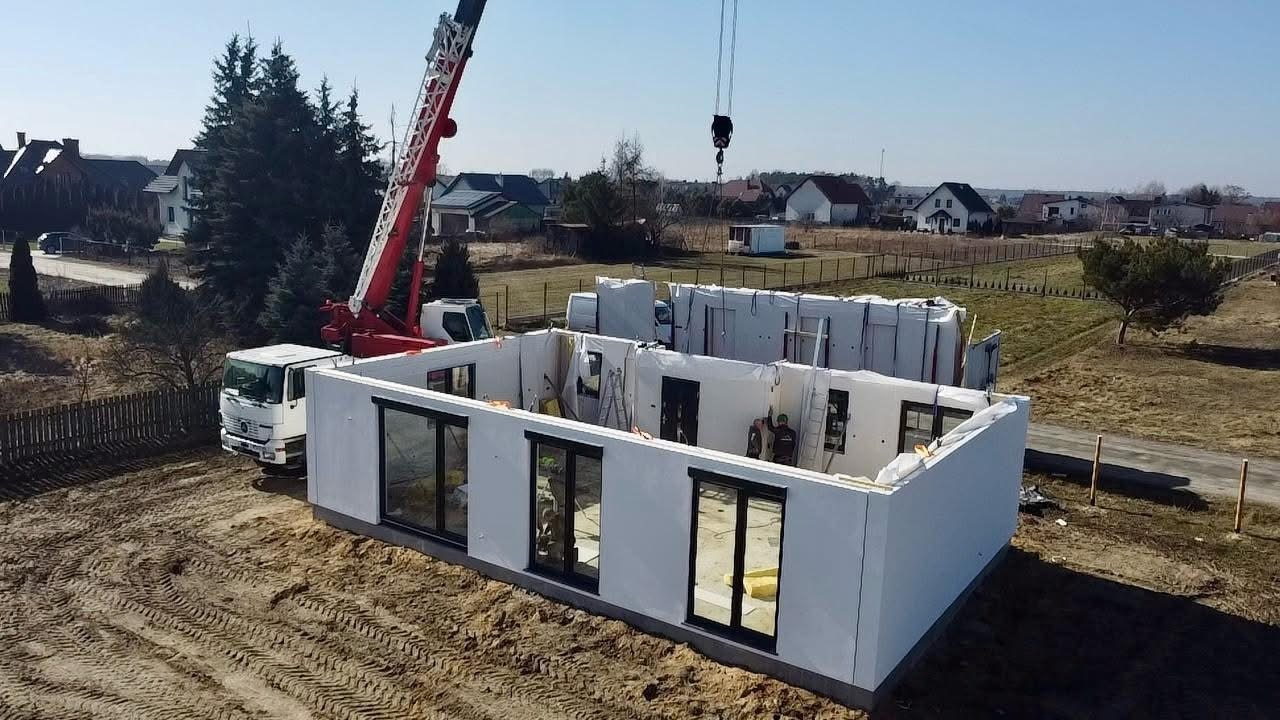 Montaż domu modułowego z użyciem dźwigu. Widoczne elementy konstrukcyjne, okna i pracownik wewnątrz. W tle zabudowa jednorodzinna i drzewa.