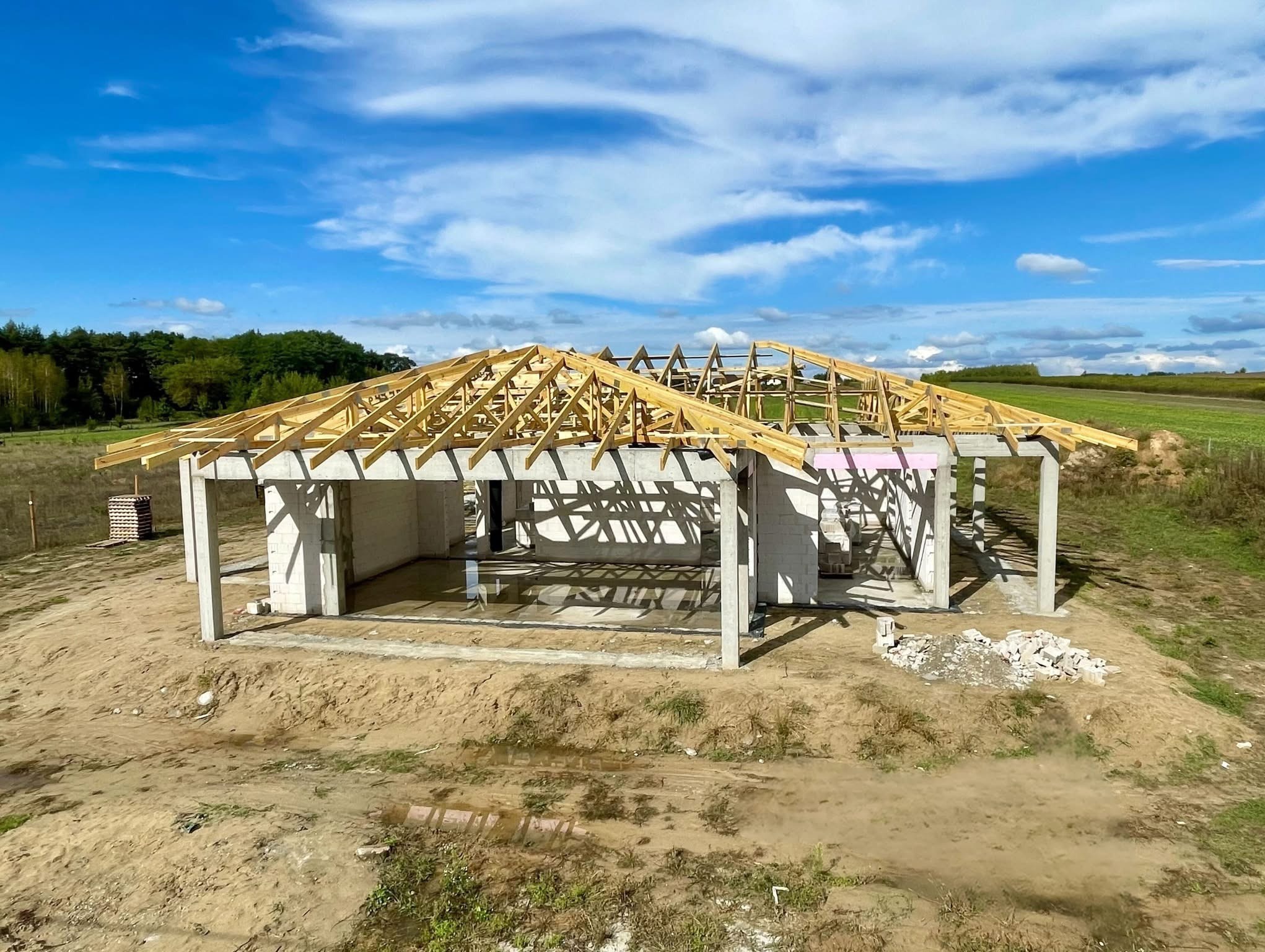 Budowa domu murowanego w stanie surowym otwartym z drewnianą konstrukcją dachu na tle błękitnego nieba. Widoczne pustaki i elementy konstrukcyjne.