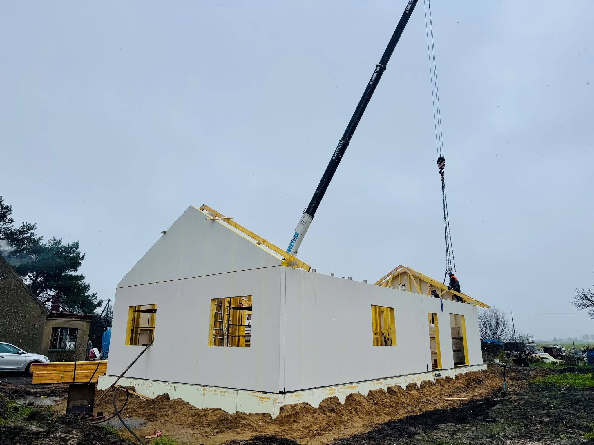 Montaż domu modułowego z użyciem dźwigu. Białe ściany z otworami okiennymi, drewniana konstrukcja dachu w trakcie budowy. Na zewnątrz widoczny krajobraz.