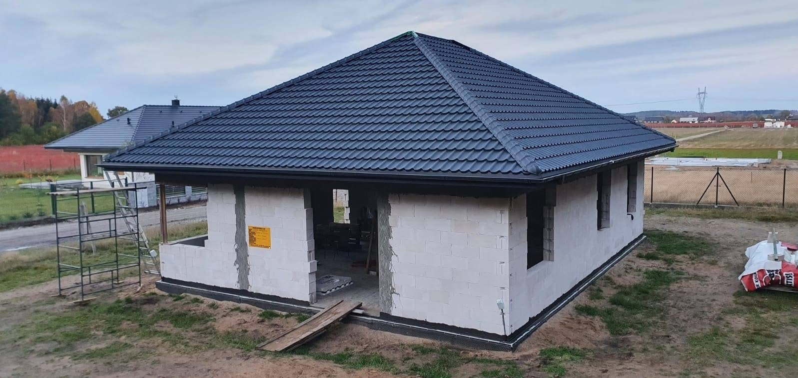 Budowa domu jednorodzinnego z ciemnym dachem z blachodachówki, widoczne białe ściany z pustaków i rusztowanie z boku. W tle krajobraz wiejski i inny dom.